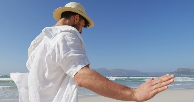 Man wearing hat and white shirt walking on sandy beach with ocean waves crashing. Perfect for promoting travel destinations, summer vacations, beachwear, and lifestyle blogs. Captures tranquility, freedom, and relaxation.