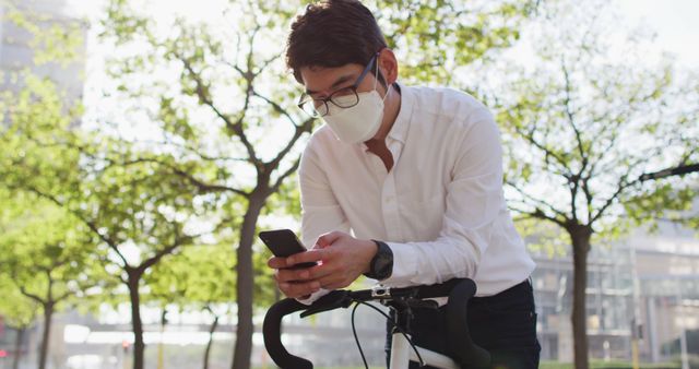 Businessman standing outdoors in park, looking at smartphone while wearing mask, with bicycle in foreground. Ideal for depicting mobile work, social distancing practices, outdoor recreation, health precautions, and modern urban lifestyle.