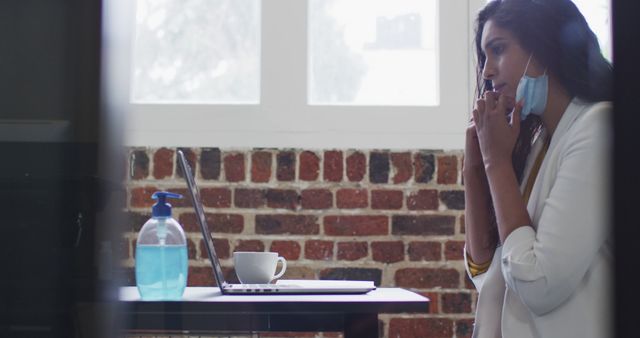 A businesswoman in casual office attire talks on the phone while sitting at her desk, concerned expression. She wears and partially removes a face mask, reflecting the pandemic's impact on the workplace. A bottle of hand sanitizer sits on the desk. Perfect for illustrating themes related to remote work, pandemic effects, workplace stress, or health and safety measures.