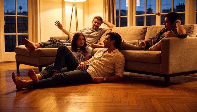 Four friends lounging and chatting on sofa at dusk in cozy living room with warm lamp glow