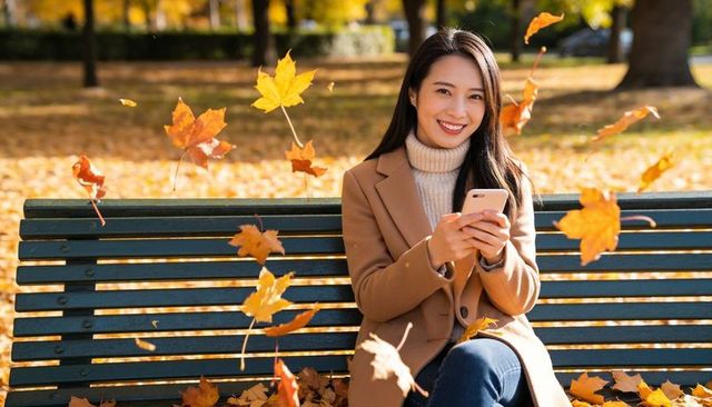 Smiling asian woman texting on park bench in camel coat and turtleneck amid autumn leaves