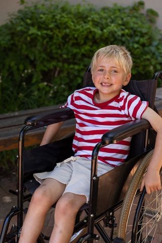 Smiling blond boy sitting in wheelchair on sunny park walkway with bench and greenery