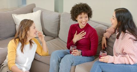 Diverse women laughing and chatting on sofa while holding drinks