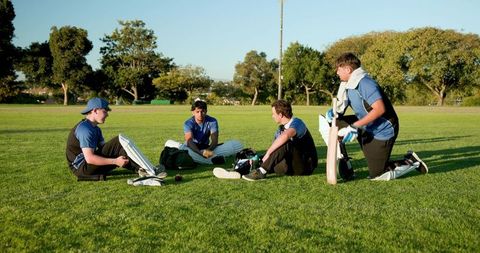 Multicultural cricket teammates resting on grass with bat, pads and gear during practice