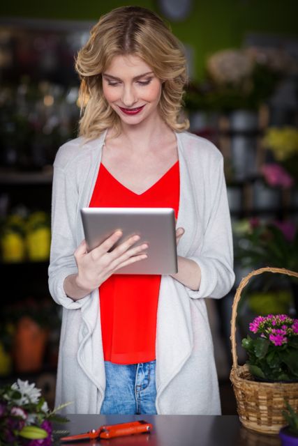 A happy female florist is using a digital tablet in a flower shop. She is smiling and appears to be engaged with her work. The setting includes various flowers and a basket, indicating a retail environment. This image can be used for promoting small businesses, technology in retail, or customer service in floral shops.