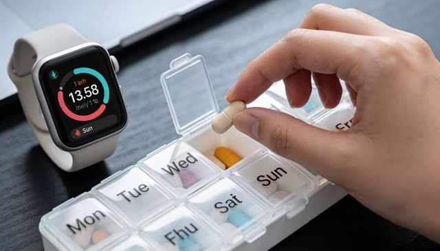 Holding hand placing daily pill into weekly pill organizer next to smartwatch display