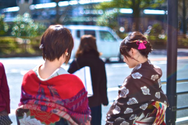 This image shows two women dressed in traditional Japanese kimono, standing and engaging in a conversation on an urban street on an autumn day. The vibrant colors of their clothing stand out against the blurry city background. Ideal for content related to Japanese culture, fashion, urban settings, and authenticity in traditional wear.