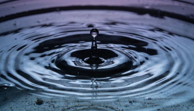 Macro rising water droplet forming column with concentric ripples under cool blue light