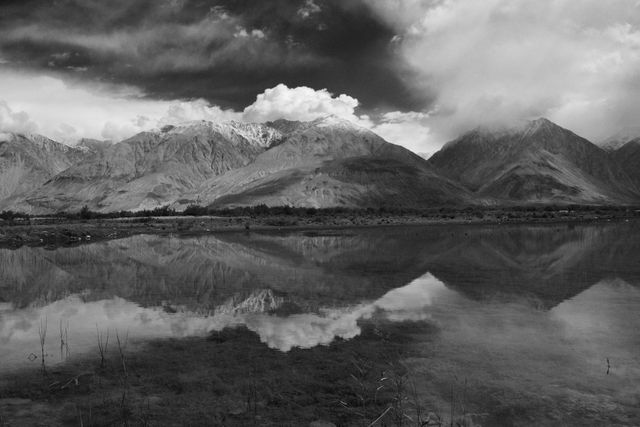 This photographic image captures the dramatic scenery of snow-capped mountains mirrored in a calm lake, under stormy clouds. The striking contrast and monochrome tones enhance the serene yet imposing atmosphere of the wilderness. Ideal for use in nature photography, travel blogs, environmental campaigns, or artistic prints, this image evokes feelings of awe and peace in the face of nature's grandeur.