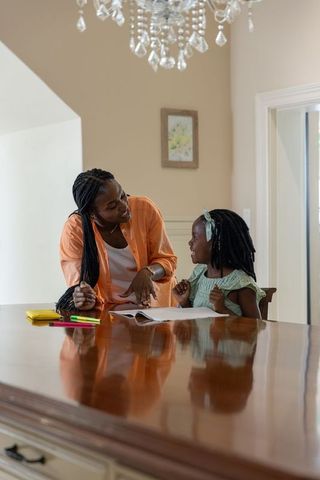 African american mother tutoring daughter at dining table doing homework and smiling
