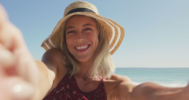 Young woman wearing straw hat, taking selfie at the beach, enjoying sunny day. Suitable for travel blogs, vacation ads, summer fashion promotions, and lifestyle content.