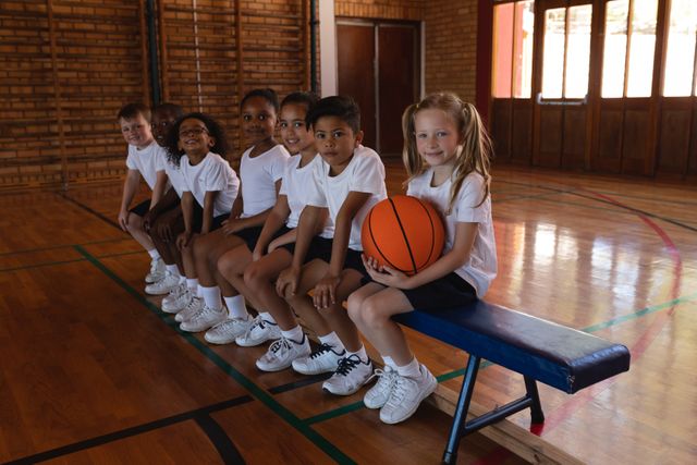 Group of schoolchildren in sports uniforms sitting on a bench in a gymnasium, holding a basketball and smiling at the camera. Ideal for use in educational materials, sports promotions, team-building activities, and diversity representation.