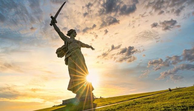 Backlit monumental heroine statue standing with sword on grassy hill during golden hour sunburst