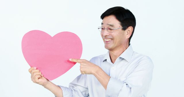 An Asian middle-aged man is smiling and pointing at a large pink heart he's holding, with copy space. His cheerful expression suggests a theme of love, affection, or Valentine's Day celebration.