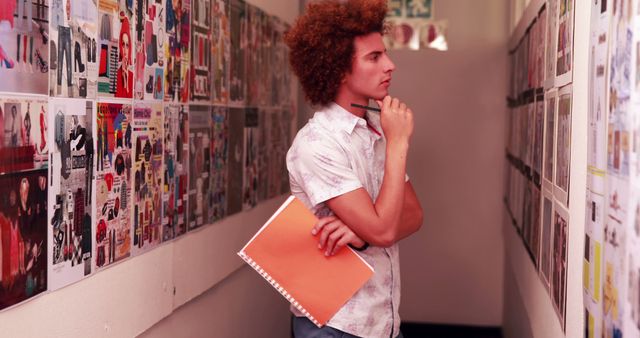 Young man with afro hairstyle analyzing a mood board in an office hallway, holding an orange notebook and pen. Ideal for use in articles about creative professions, brainstorming sessions, fashion design, and interior decoration.