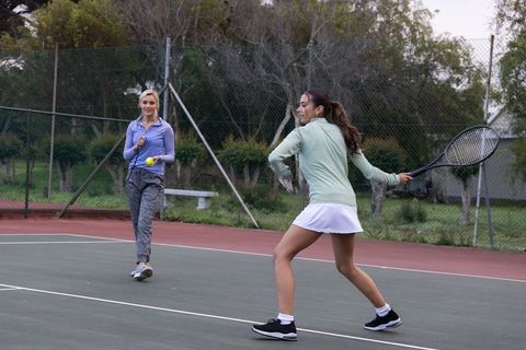 Women practicing tennis on outdoor court during coaching drill, swinging racket and tossing ball