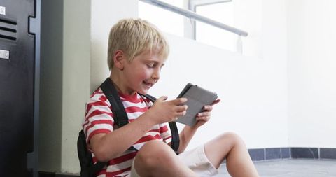 Smiling blond schoolboy sitting in hallway using tablet while leaning against locker pillar