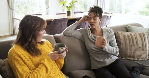 Multiracial women sharing coffee and chatting on cozy sunlit living room sectional sofa