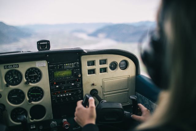 Pilot's hands on aircraft controls in cockpit, showing instrumentation and mountains in distance. Ideal for travel and adventure themes, aviation industry material, pilot training illustrations, or blogs about flying and aeronautics.