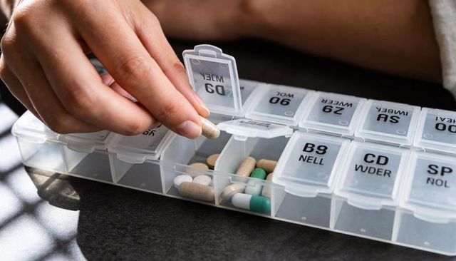Adult hand holding beige capsule from labeled weekly pill organizer with white robe sleeve