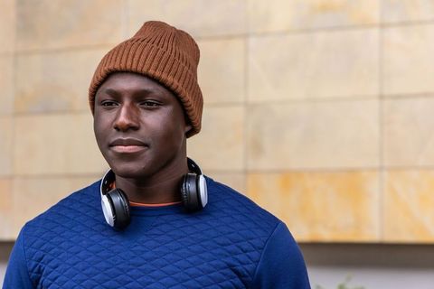 Young man wearing brown beanie and headphones standing against beige tile facade in navy pullover