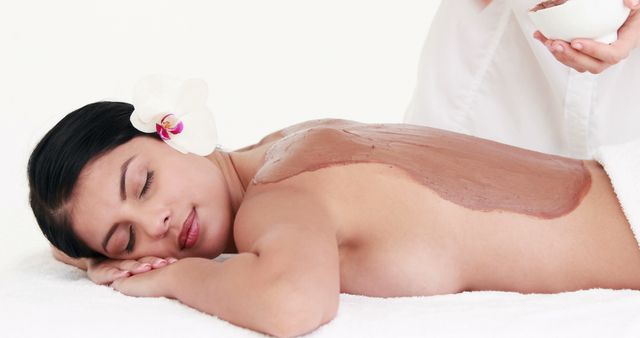 Image depicting a woman receiving a luxurious chocolate body mask treatment. With an orchid in her hair, she is lying on a white towel and appears relaxed and comfortable. Ideal for promoting spa services, beauty products, wellness blogs, or relaxation therapy articles.