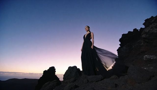 Gazing woman standing on rocky outcrop wearing flowing black gown at twilight