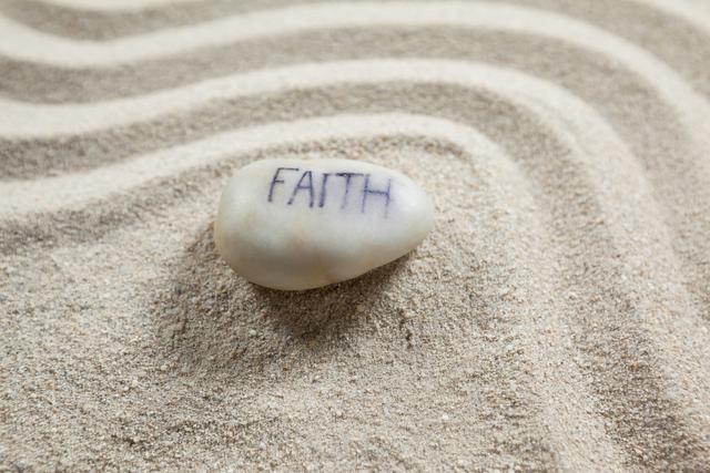 White pebble with the word 'faith' engraved, placed on sand with wavy patterns. Ideal for use in themes related to spirituality, meditation, inspiration, and tranquility. Suitable for blogs, social media posts, and websites focusing on mindfulness and inner peace.
