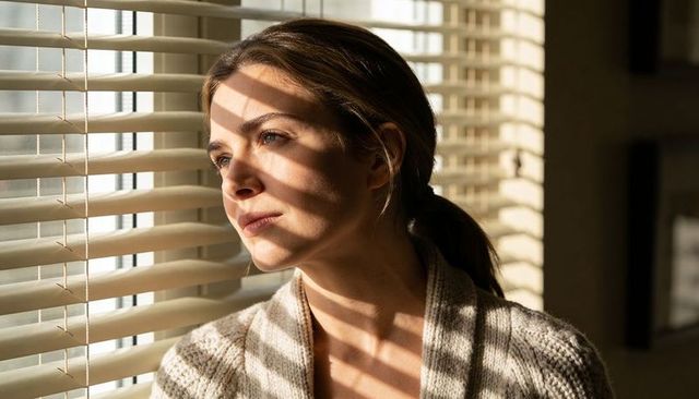 Woman gazing through blinds with sunbeam stripes on face and cardigan