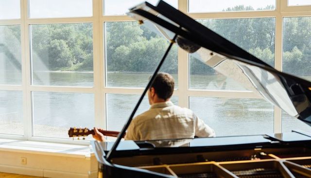 Sitting musician holding acoustic guitar beside grand piano gazing out river view in light-filled ro