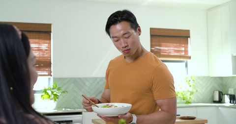 Young man mixing fresh salad with chopsticks at bright modern kitchen island, casual couple