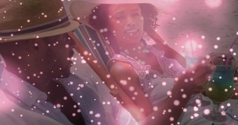 Couple relaxing on striped beach chairs sipping teal cocktails wearing sun hats with pink bokeh