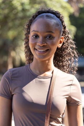 Sunlit african woman smiling in park wearing taupe tee and crossbody bag