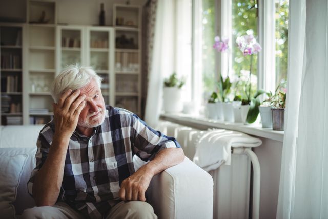 Elderly man sitting on a sofa in a living room, looking worried and thoughtful. He is wearing a plaid shirt and is surrounded by indoor plants near a window with natural light. This image can be used to depict themes of aging, mental health, loneliness, and contemplation. Suitable for articles, blogs, and advertisements related to senior care, mental wellness, and home living.