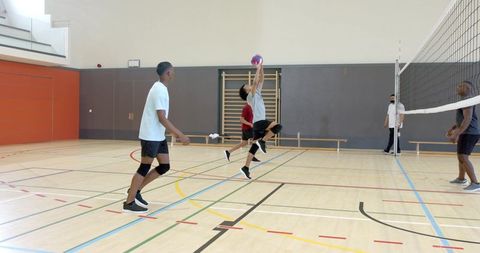 Diverse male volleyball team jumping and reaching for ball at indoor gym net daylight play