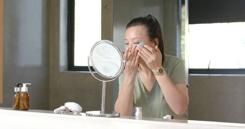 Asian woman applying under-eye patches at bathroom vanity during morning skincare routine