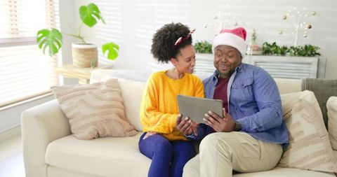 Diverse couple sharing holiday laughter while watching tablet on cozy sofa with Santa hat
