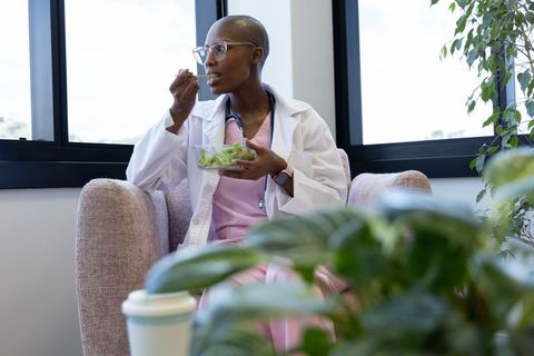 African woman doctor eating salad in clinic lounge on break wearing scrubs and lab coat