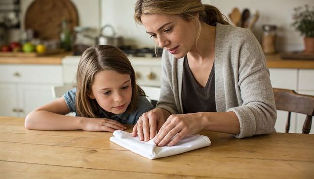 Mother teaching daughter folding napkin on wooden kitchen table, cozy family moment