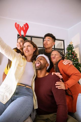 Diverse friends taking festive selfie by christmas tree indoors