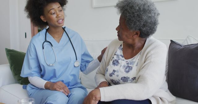 Nurse dressed in blue scrubs engaging in compassionate conversation with an elderly woman sitting on a sofa. Elderly woman is wearing a white cardigan over a floral blouse. Scene represents importance of in-home healthcare, medical support, patient comfort, and caregiver relationships. This can be used for healthcare, caregiving campaigns, senior living advertisements, or medical services marketing.