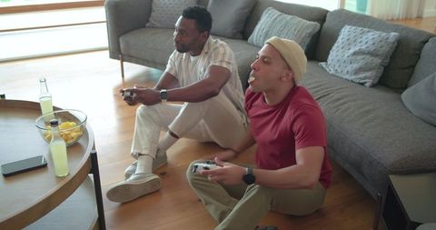 Diverse friends playing video games on living room floor, snacking and relaxing