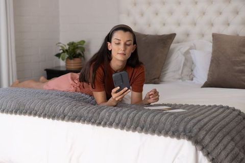 Woman reclining on bed using smartphone in serene neutral bedroom with ribbed grey throw