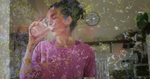 Mature woman drinking water from glass in kitchen with foliage double exposure overlay