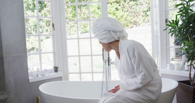 A woman in a white bathrobe and hair towel sits by a large bathtub in a modern bathroom with big windows. The bright setting and green garden outside create a serene and calming environment. Perfect for wellness, self-care, and relaxation themes. Suitable for advertisements related to home decor, bath products, or spa services.