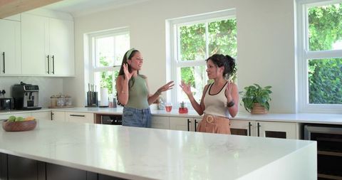 Two women conversing and gesturing in bright modern kitchen with marble island