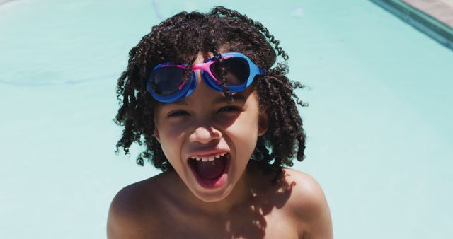 Young child with curly hair wears swimming goggles while playing at a swimming pool on a bright sunny day. The open-mouthed expression shows excitement and joy, making it ideal for promoting children's activities, summer camps, pool safety tips, and leisure products aimed at families. This image captures the essence of childhood fun and energy.