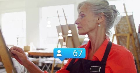 Mature artist painting at easel while receiving social media followers notification