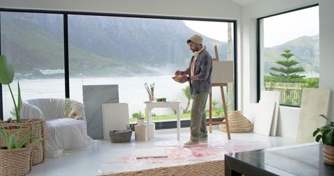 Indian artist mixing paint at easel in waterfront studio with mountain view and natural light