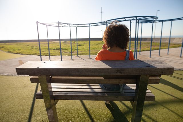 This image shows a woman sitting on a bench at a seaside playground on a sunny day. Ideal for use in lifestyle blogs, articles about relaxation and leisure, travel websites, or promotional materials for parks and recreational areas.